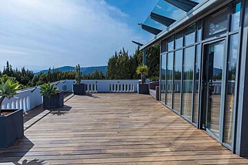 A spacious wooden deck terrace featuring a modern glass-paneled building, white balustrade, potted plants, and a scenic view of green hills.