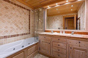A rustic bathroom featuring light wood cabinets, a double vanity, a large mirror, and a bathtub surrounded by beige tiles.