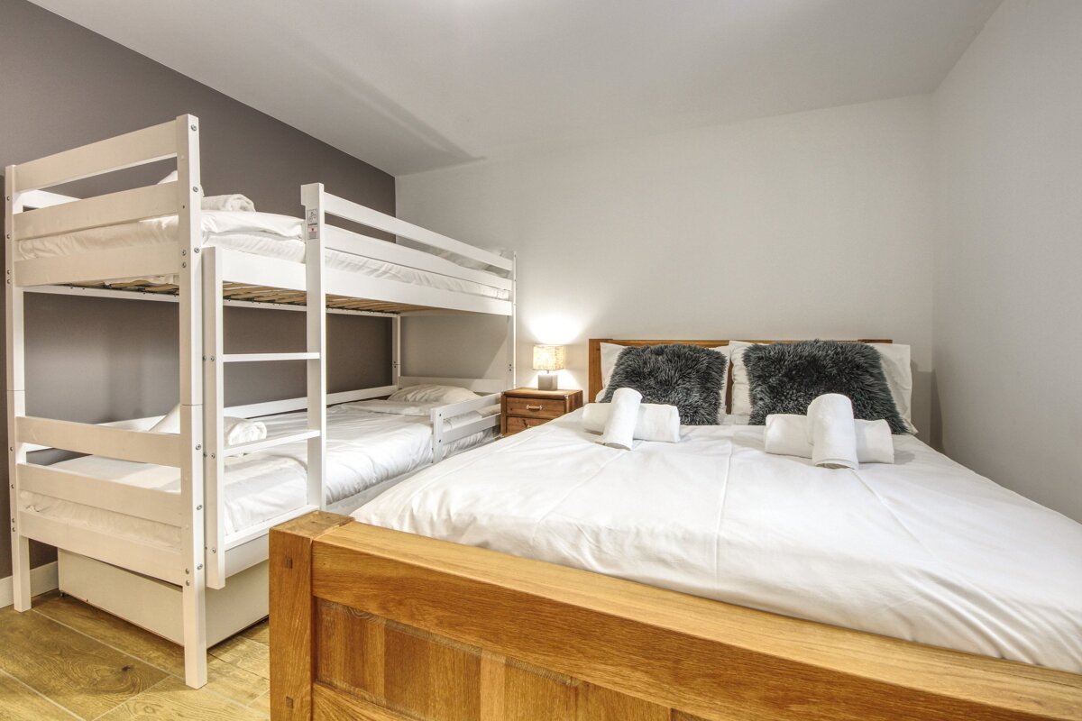 A bright bedroom features a double bed with white bedding and fluffy pillows, next to white bunk beds and a pull-out trundle bed, on a wooden floor.