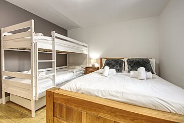 A bright bedroom features a double bed with white bedding and fluffy pillows, next to white bunk beds and a pull-out trundle bed, on a wooden floor.