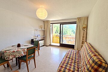 A cozy room features a floral-covered dining table, plaid sofa, and balcony with mountain and tree views. A paper lantern illuminates the space.