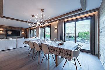 Modern chalet dining room with a long table, white chairs, branching chandeliers, and glass sliding doors opening to a mountain view.