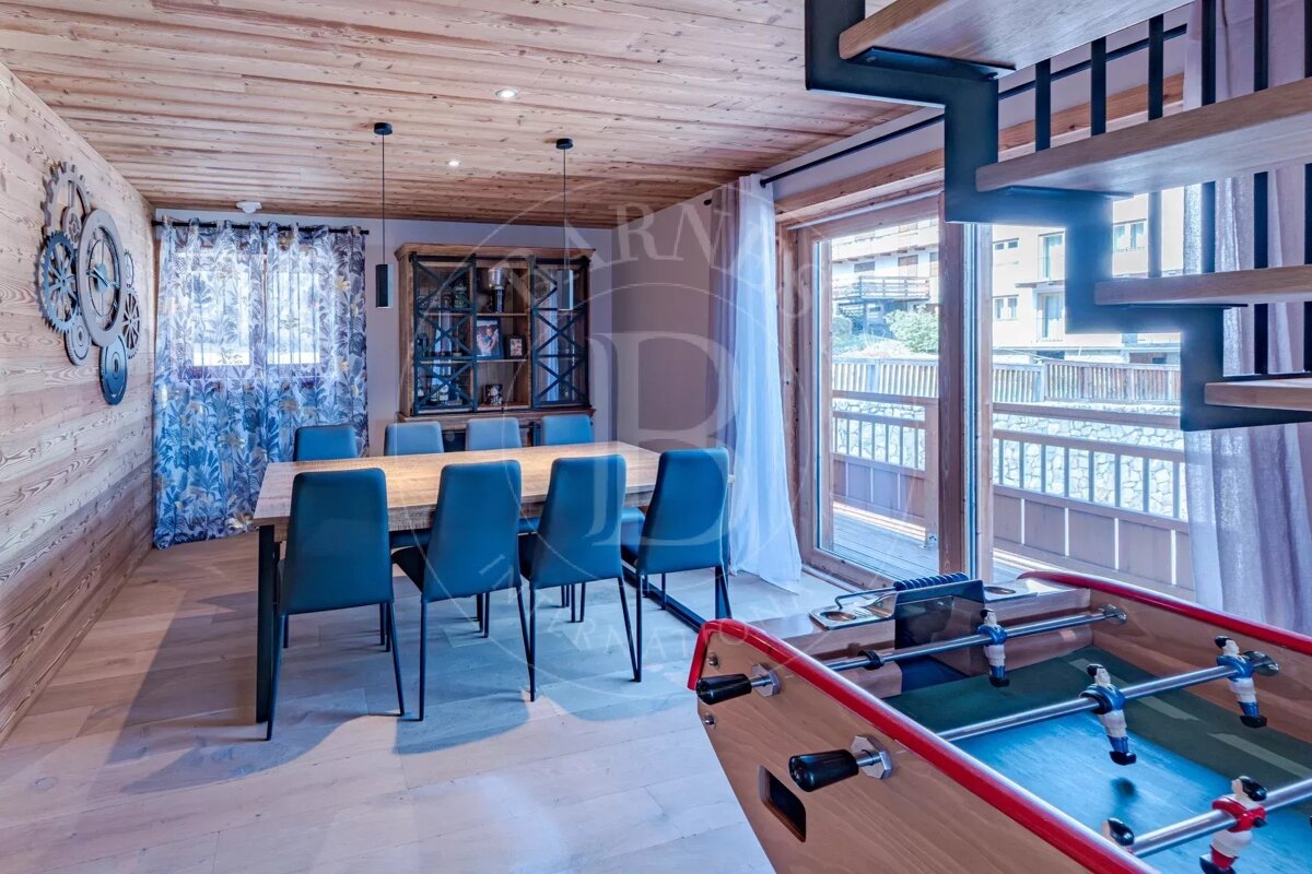 A cozy chalet dining area with blue chairs, a wood table, a foosball table in the foreground, and an open staircase.