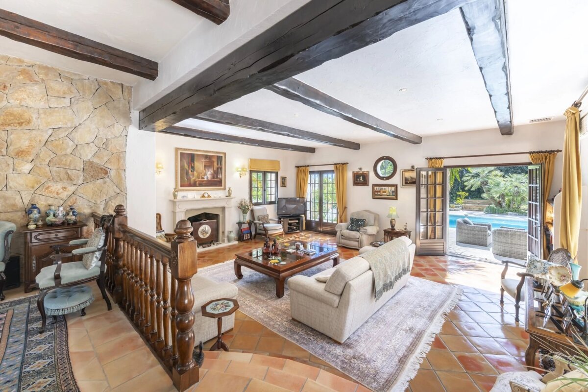 Spacious living room with exposed beams, stone wall, terracotta floor, and open doors revealing a pool and lush garden.