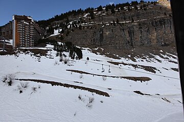 A snowy mountain resort with large buildings, a rocky cliff, and ski lift chairs traversing the slopes under a clear sky.