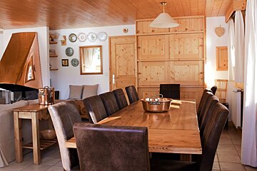 A sunny, rustic dining room. Features include a long wooden table, brown leather chairs, a copper pot, and a copper fireplace in a wood-paneled space.