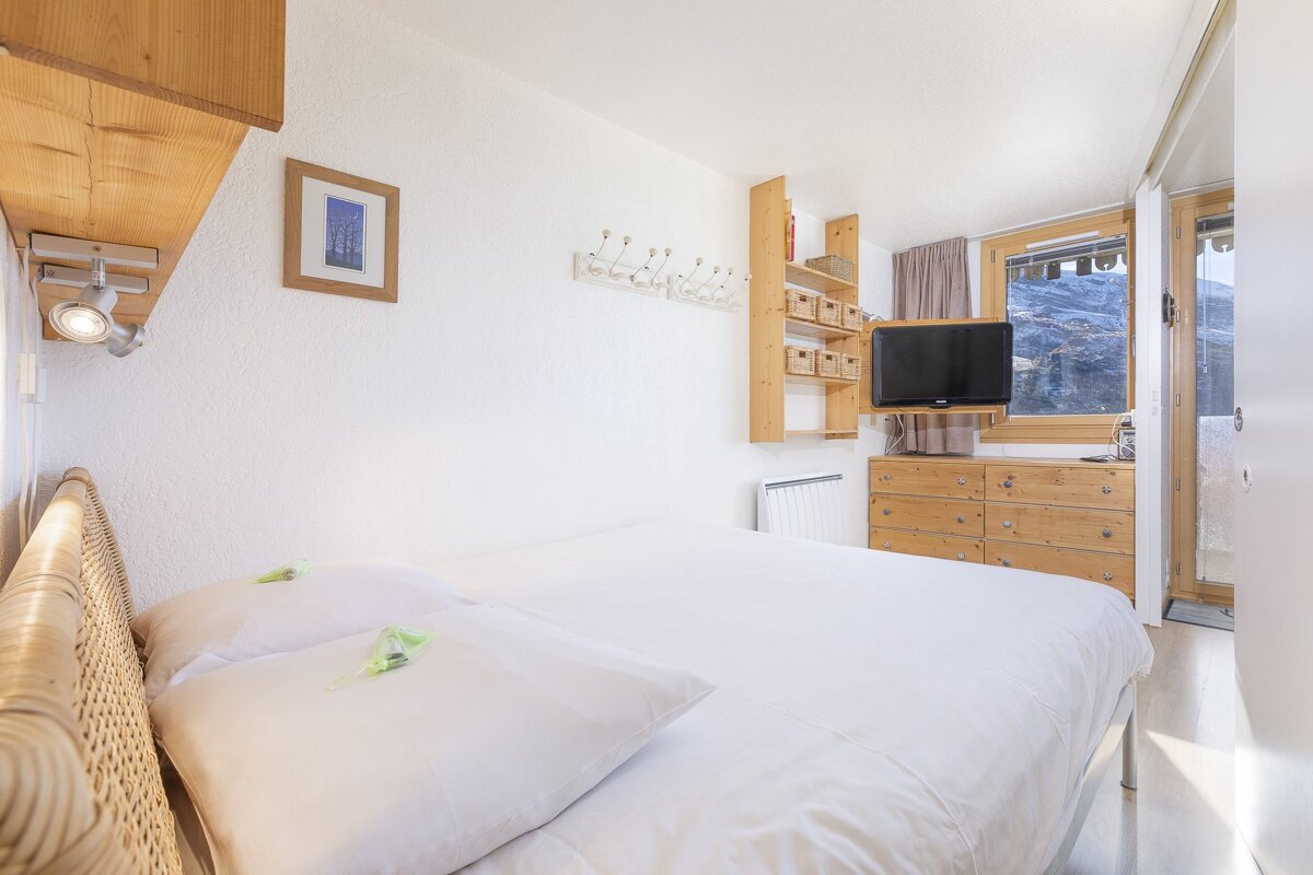 A cozy, light-filled bedroom with a white bed, wooden furniture, and a TV. A large window offers a stunning mountain view.