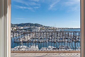 Balcony view of a sunny marina packed with white boats, a coastal town, and green hills under a bright blue sky.