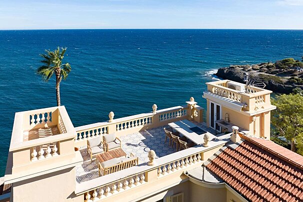Luxurious coastal villa terrace with elegant balustrades, outdoor dining/lounge areas, a tall palm tree, and a breathtaking blue ocean view.