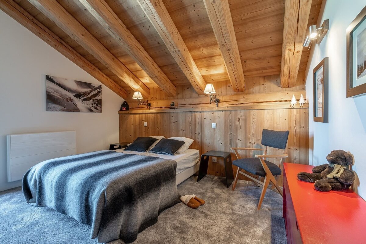 Rustic, cozy attic bedroom with exposed wooden beams, wood paneling, a bed, chair, and a teddy bear by a red chest.