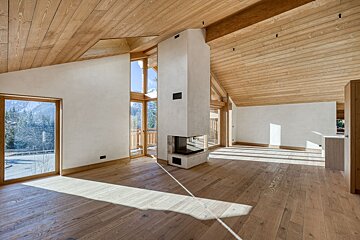 An empty, modern wooden chalet interior with a central white fireplace, sloped ceilings, and large windows letting in bright sunlight.