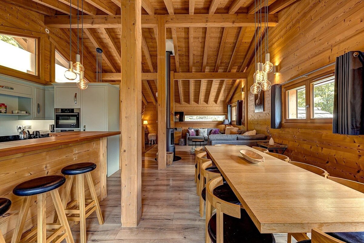 A warm, open-plan wooden chalet interior featuring a modern kitchen, long dining table, and cozy living area. Exposed beams and contemporary lighting create a stylish feel.