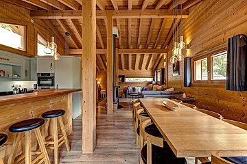 A warm, open-plan wooden chalet interior featuring a modern kitchen, long dining table, and cozy living area. Exposed beams and contemporary lighting create a stylish feel.