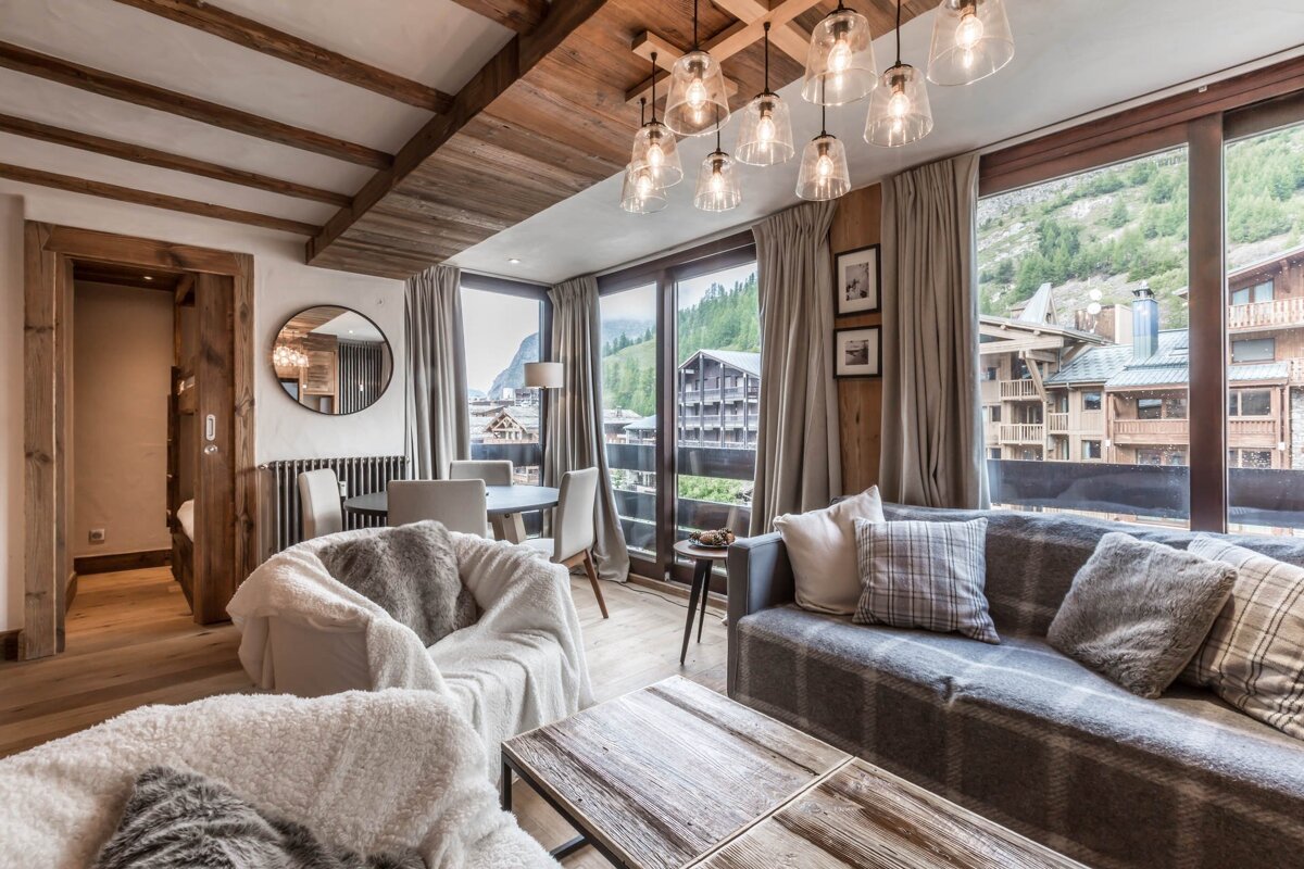 A cozy, rustic living room with exposed wood beams, a plaid sofa, fluffy armchairs, and large windows offering a view of a mountain village.