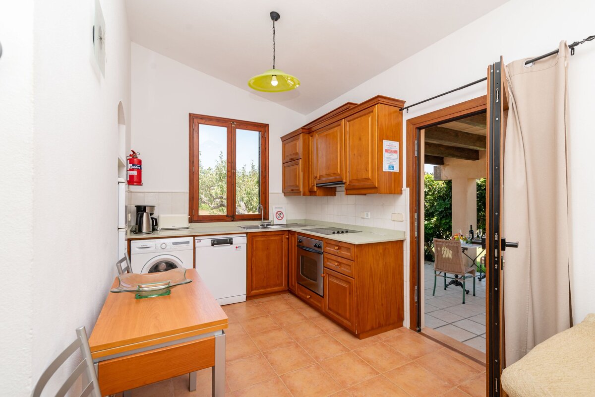 A bright kitchen features warm wooden cabinets, integrated appliances, and a dining table. A window overlooks trees, and a door opens to an outdoor patio.