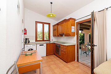 A bright kitchen features warm wooden cabinets, integrated appliances, and a dining table. A window overlooks trees, and a door opens to an outdoor patio.