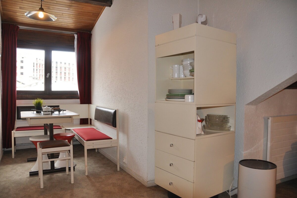 A cozy kitchen/dining area with a wooden ceiling, window with red curtains, a small table, benches with red cushions, and a cabinet full of dishes.