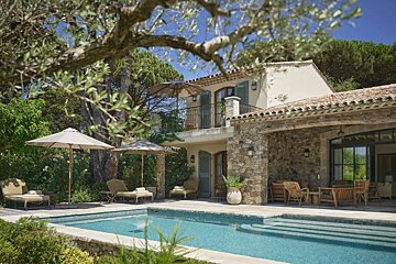 A serene Mediterranean villa featuring a swimming pool, lounge chairs with umbrellas, and lush greenery, bathed in sunlight.