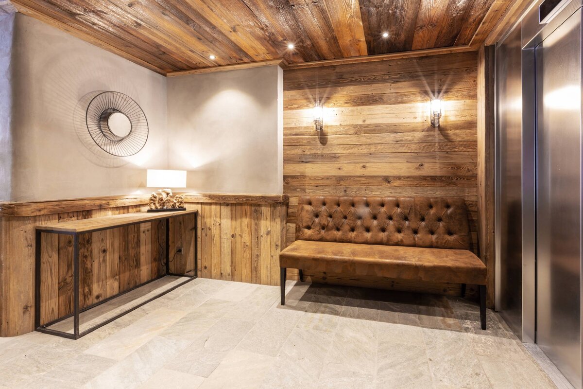 Cozy chalet-style foyer featuring rustic wood walls/ceiling, a tufted leather bench, modern console, and elevator.
