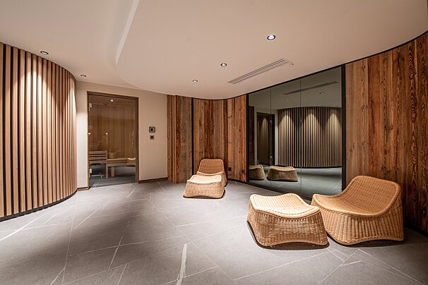 A modern spa relaxation area featuring curved wooden walls, wicker seating, a large mirror reflecting slatted wood, and a glass-doored sauna.