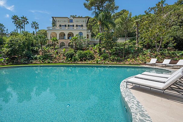 A luxury Mediterranean villa with a curved turquoise swimming pool, loungers, and lush tropical gardens under a clear blue sky.