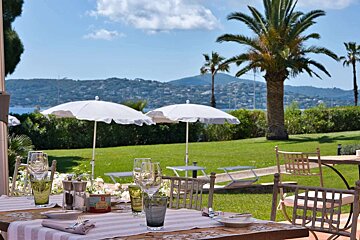 Outdoor dining table set on a patio, overlooking a lush green lawn, white umbrellas, palm trees, and a scenic bay with distant hills.