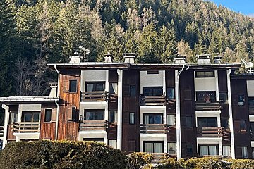 Alpine-style apartment building with wooden balconies set against a lush pine forest on a hillside.