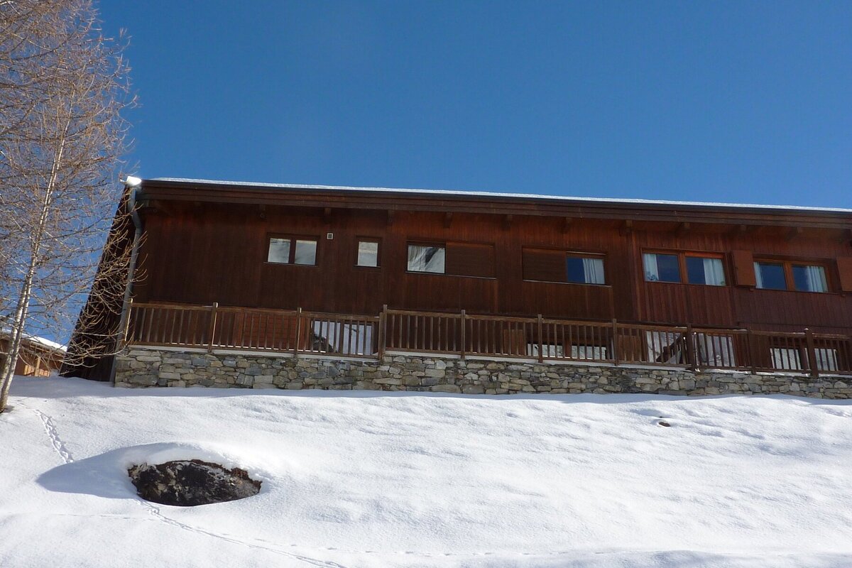 A wooden chalet with a stone foundation stands on a snow-covered hill under a clear blue sky.