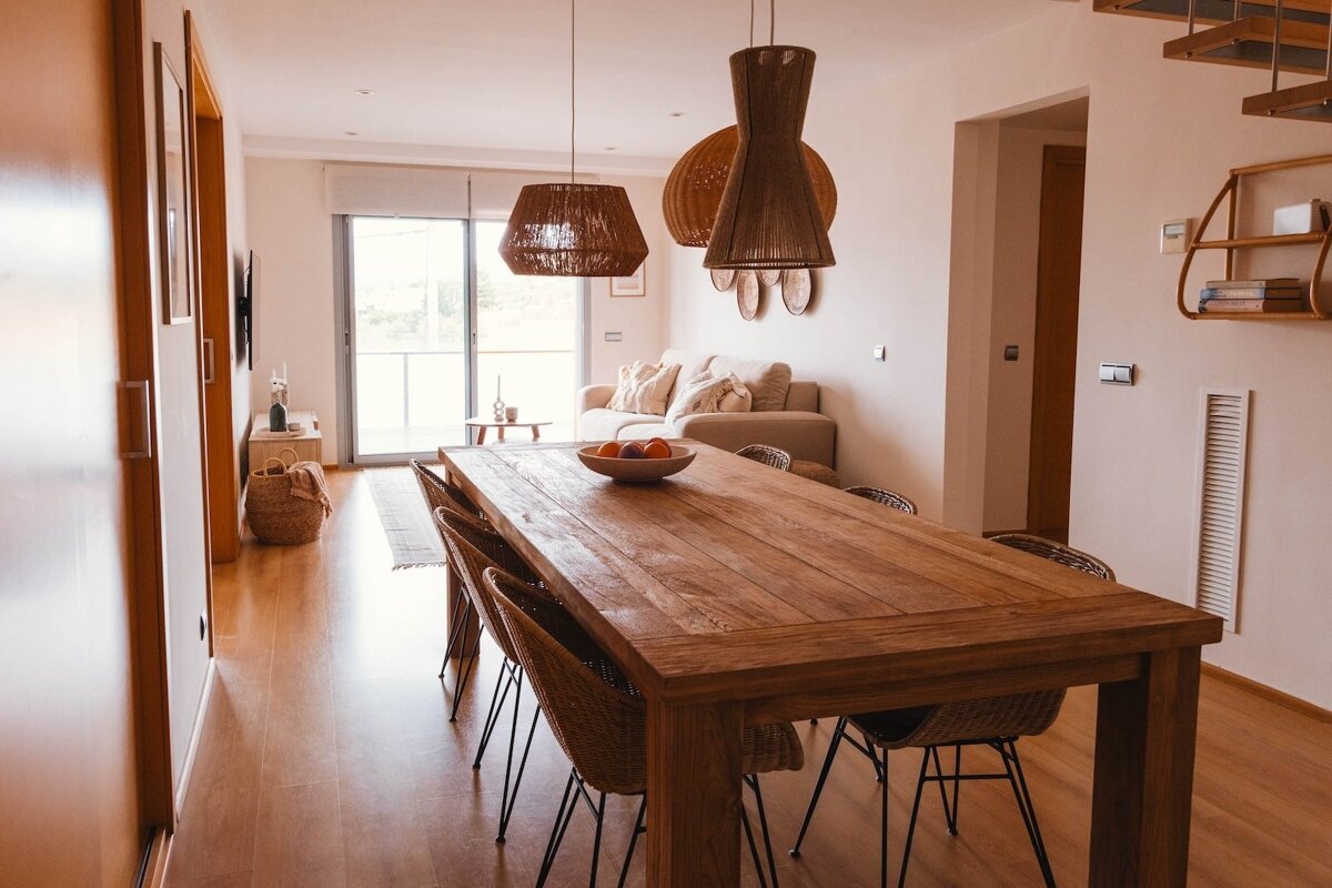 Warm, inviting open-plan dining and living space with a long wooden table, wicker chairs, woven pendant lights, and earthy decor.