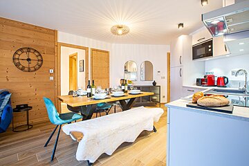 A cozy open-plan kitchen and dining room with wooden walls, a set dining table, a furry bench, and a modern kitchen with red accents.