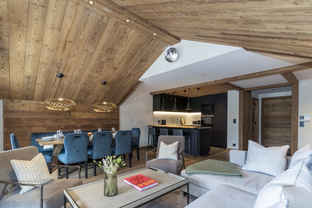 A spacious, modern chalet interior with rich wooden ceilings, a dark kitchen island, a dining area with blue velvet chairs, and a cozy living space.