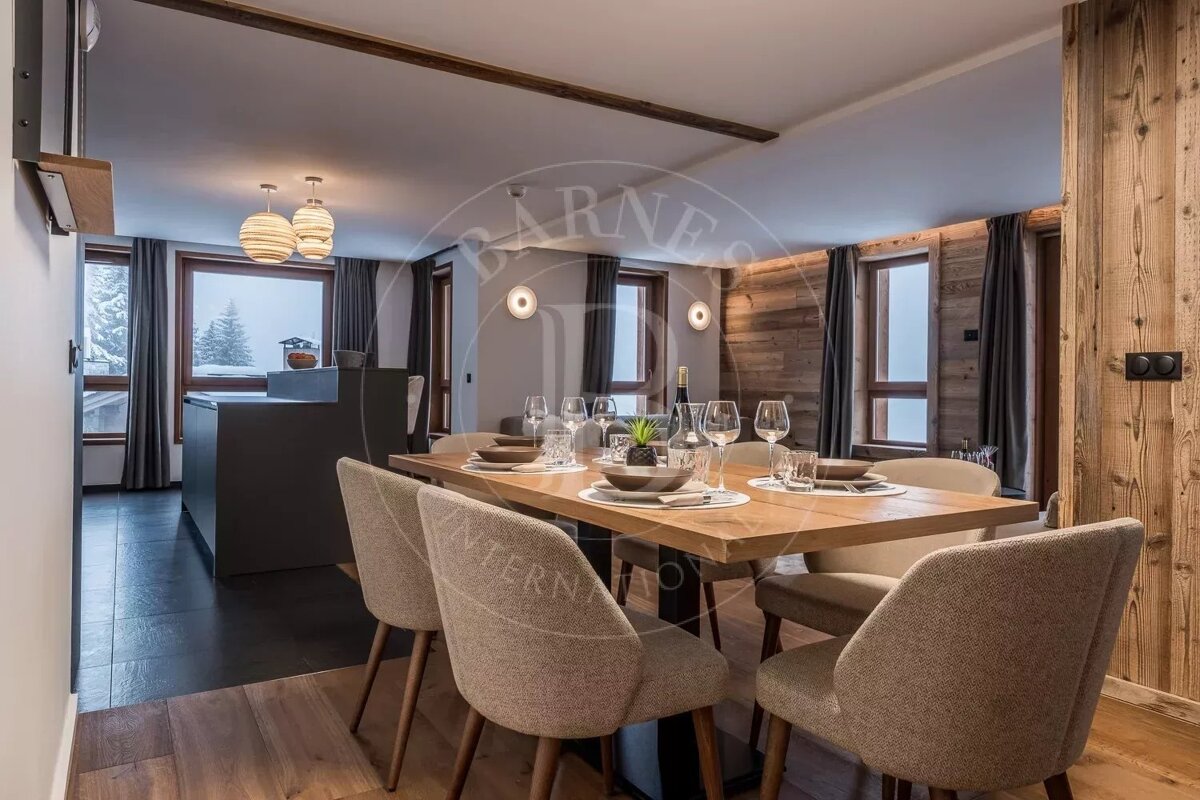 A modern chalet dining room with a wooden table set for dinner, connected to a kitchen, featuring wood-paneled walls and snowy mountain views.