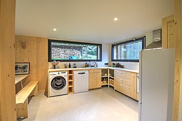 Cozy, rustic kitchen with light wood cabinets, washer, dishwasher, and a dining bench. Windows show stacked firewood outside.
