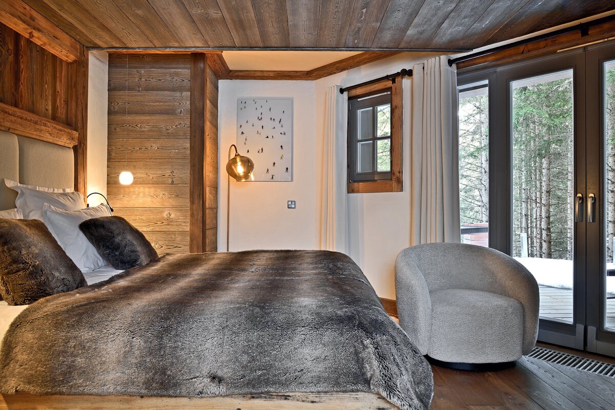 Cozy, rustic bedroom featuring wood paneling, a large bed with a faux fur throw, and a glass door opening to a snowy forest view.