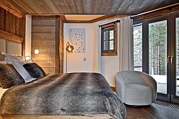 Cozy bedroom with a fur-covered bed, wooden walls & ceiling, and large glass doors revealing a snowy forest. A warm, inviting atmosphere.