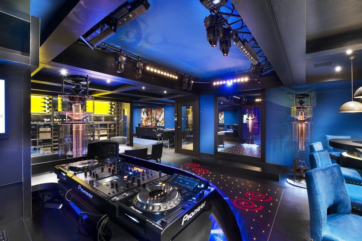 Luxurious home nightclub with a DJ setup, blue lighting, a lit dance floor, and a wine display, all set for an energetic party.