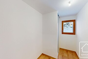 An empty room with white walls, wooden floor, and a window framing a snowy outdoor scene. A ceiling light illuminates the space.