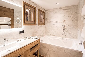 Elegant bathroom with marble walls, a wooden vanity, a bathtub, and a backlit mirror, blending modern and rustic styles.