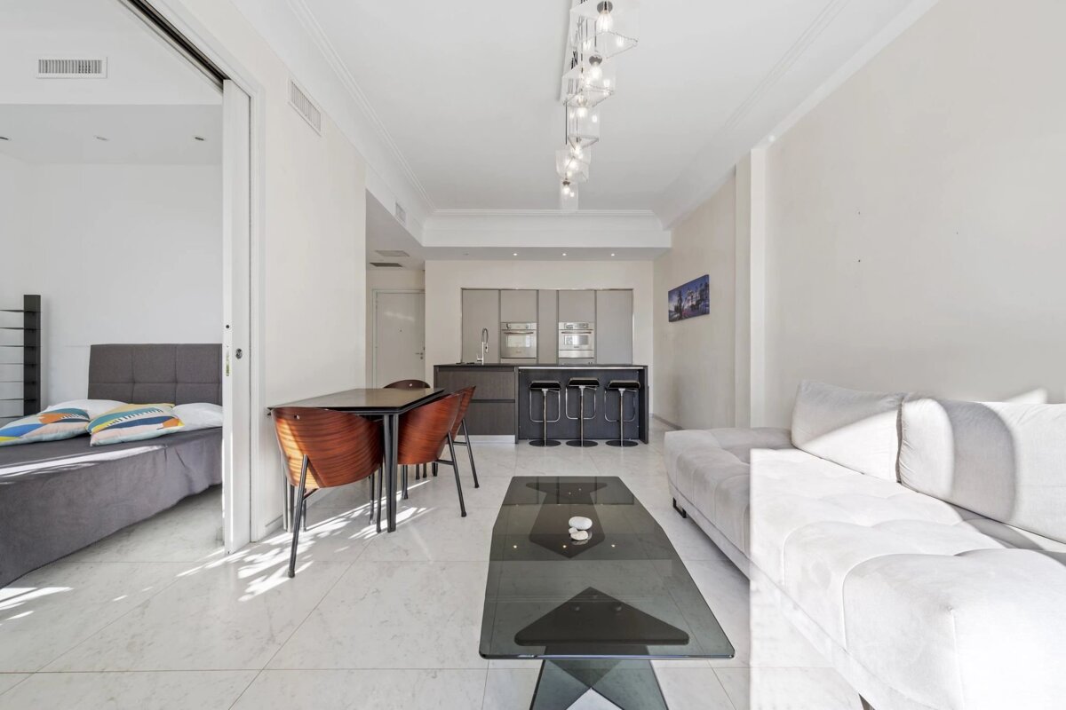 A bright, modern apartment with an open living, dining, and kitchen area, featuring a white sofa, sleek kitchen, and adjacent bedroom.