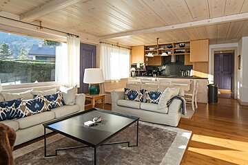 Cozy chalet living room with wood paneling, beige sofas, a black coffee table, and an open-plan kitchen in the background.