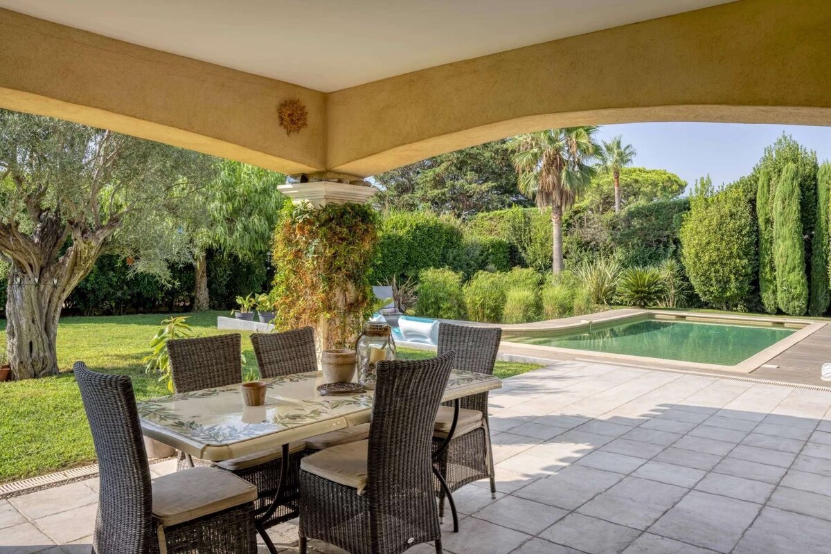 Covered patio with a dining set, overlooking a lush green garden with an olive tree, a swimming pool, and dense tropical foliage.