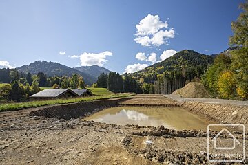 A construction site with a muddy pool and dirt piles. Beyond, two chalets on a green slope, with lush forested mountains under a clear sky.
