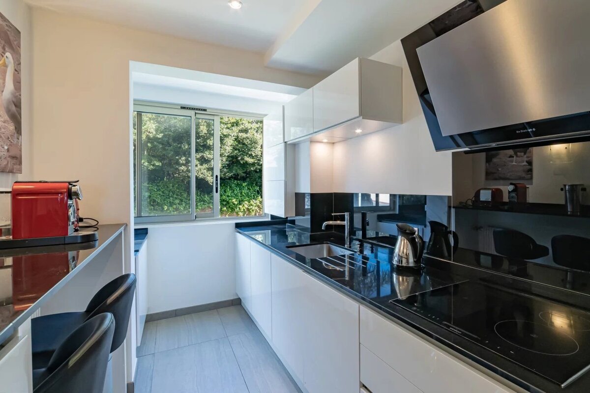 A sleek modern galley kitchen with glossy white cabinets, black countertops, a window viewing trees, and a red coffee machine.