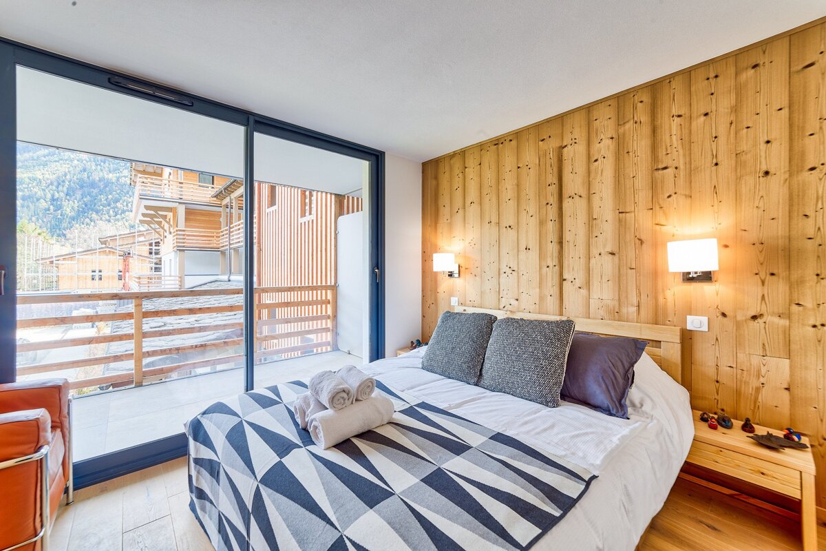A bright bedroom with a wood-paneled wall, bed with a geometric blanket, and large glass doors opening to a balcony with a view of mountain buildings.