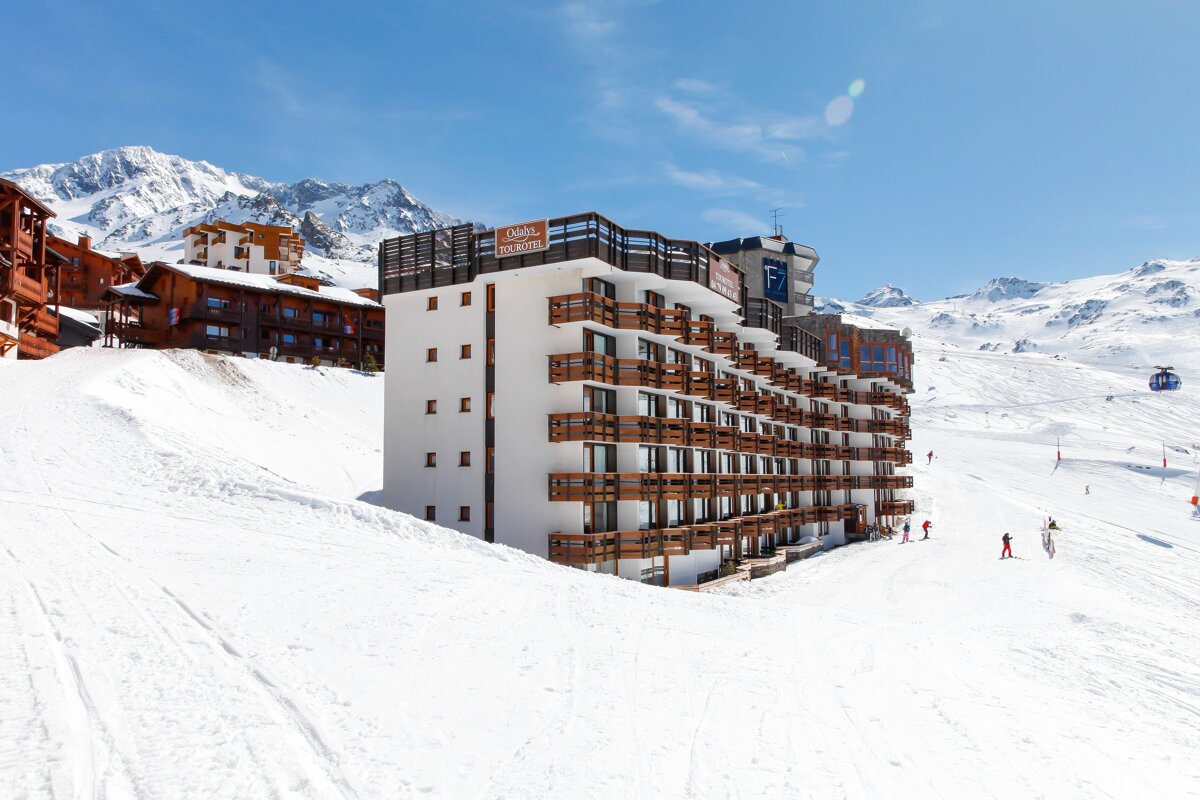 A large ski resort hotel with balconies stands on a snowy mountain slope, with skiers, a gondola, and distant peaks under a bright blue sky.