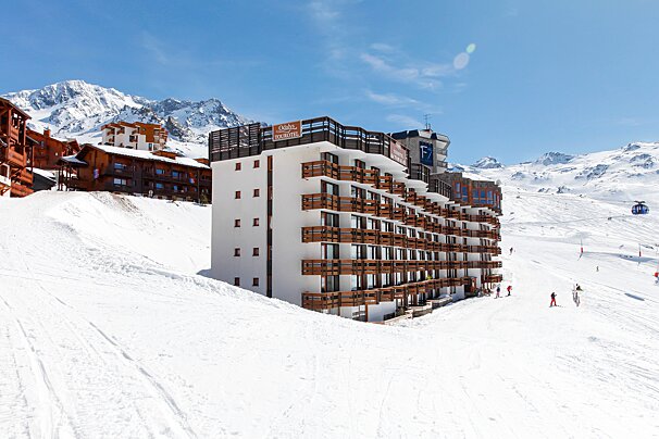 A large ski resort hotel with balconies stands on a snowy mountain slope, with skiers, a gondola, and distant peaks under a bright blue sky.