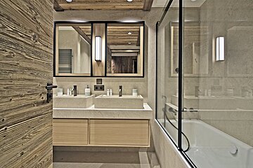 Modern chalet bathroom with double stone sinks, black-framed mirrors, a bathtub, and warm wood details on the door and ceiling.