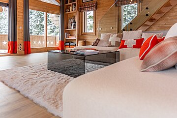 Cozy alpine chalet living room with a fluffy rug, white sofa, dark glass coffee table, and snowy views outside.