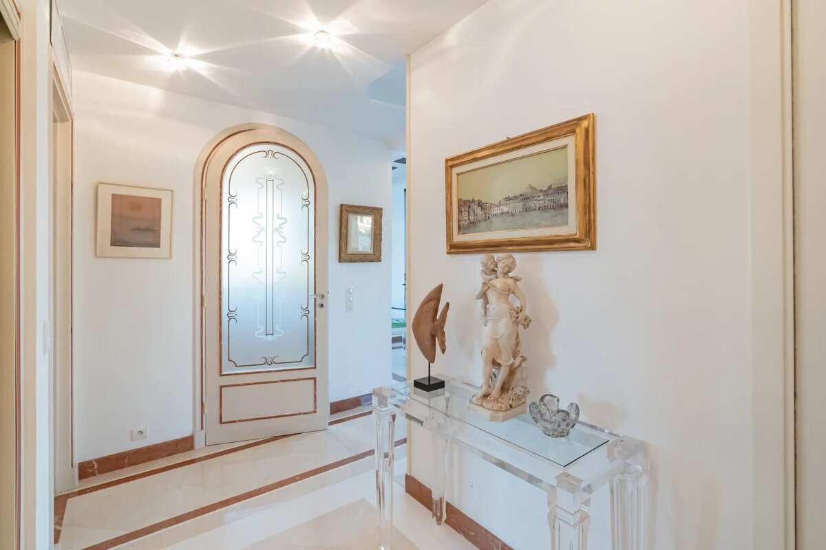 Elegant white hallway with marble floor, arched door, and a console table displaying sculptures under a framed painting.