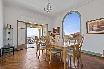 A bright dining room with a wooden table and chairs, herringbone floor, balcony with city view, and an arched window showcasing the sky.